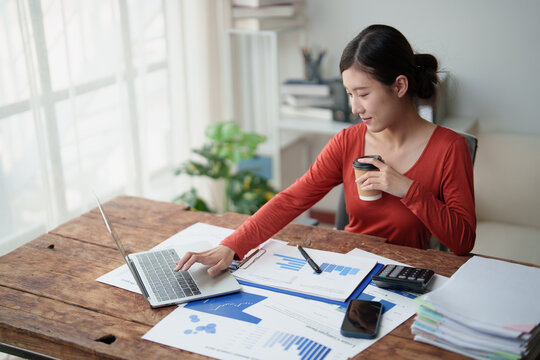 Young Asian woman focusing on work, typing on laptop, reviewing financial reports and charts, holding a coffee cup, and managing data from her home office setup