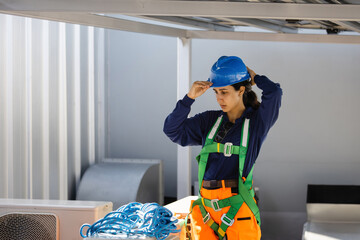 Female industrial technician putting on blue hard hat and safety harness for rooftop work,...