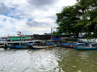 Obraz premium Small boats dock along a calm river near trees in a rural area under cloudy sky during daytime.