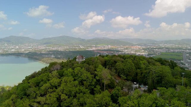 Panoramic aerial Kaokard Viewpoint Tower, a stunning vista unfolds over Phuket city, a tranquil bay, distant mountains. Lush green forest envelops the tower under bright blue sky filled white clouds