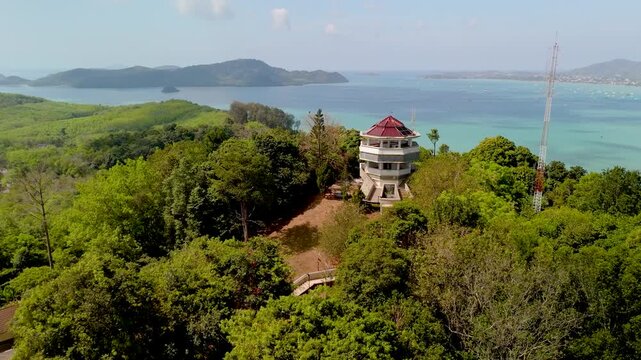 From Kaokard Viewpoint Tower, a stunning vista unfolds over Phuket city, a tranquil bay, and distant mountains. Lush green forest envelops the tower under a bright blue sky filled with white clouds