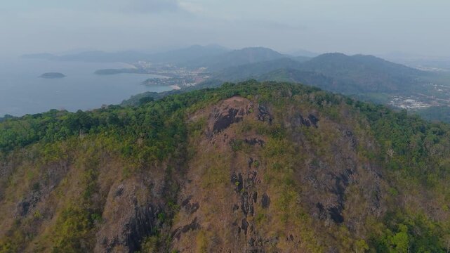 Golden sun rays fall Black Rock rugged mountains covered in green and brown foliage. A vast ocean and coastal city stretch into the distance under a dramatic orange and purple sunset sky above Phuket