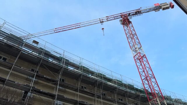 Tower crane above construction site in Germany with active facade renovation and scaffolding. Building repair, roof work and modernization in urban development under blue sky.