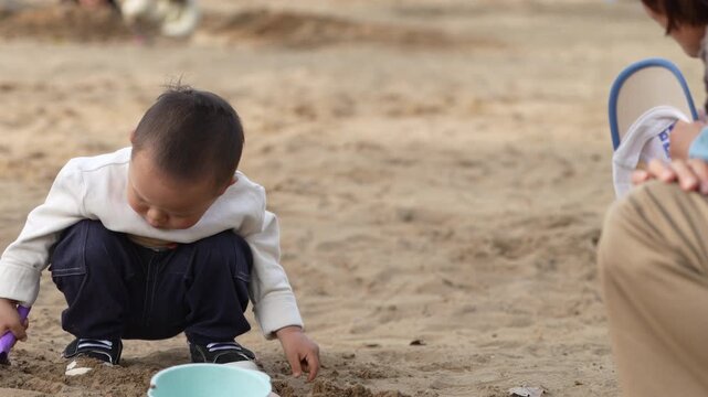 Little Asian Boy Squatting in a Sandbox and Digging Sand Outdoors.