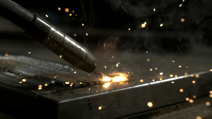 A person welds metal pieces together in a workshop. Bright sparks fly as the welding tool connects the surfaces. Smoke rises from the heated metal in dim lighting. © Videophilia