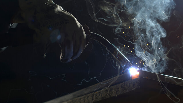 A person works on metal with a welding tool. Sparks fly and smoke rises as the metal pieces join together. The setting is a workshop where crafting and repairs happen.