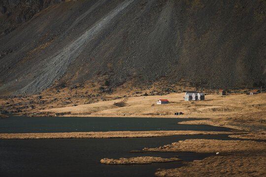 Isolated local farm buildings beneath a steep mountain in rural Iceland, Hvalnes Nature Reserve Beach, spring countryside with lake, earthy slopes, remote Nordic landscape along the Ring Road
