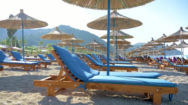 Empty sun loungers with blue cushions on a pebble beach. Rows of straw umbrellas under clear blue sky. Calm seaside resort scene, perfect for summer vacation.