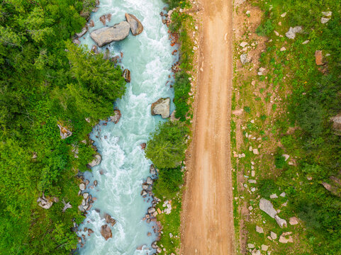 Serene mountain river flows alongside unpaved dirt road in Kyrgyzstan. Lush trees and rocks enhance the tranquil natural landscape during daytime. Directly above aerial view.
