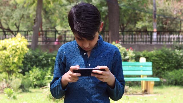 Elementary age child of Indian ethnicity standing outdoor in nature during summer season and using mobile phone.