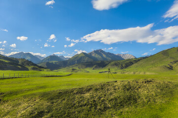 Fototapeta premium Tripod with camera equipment set against a backdrop of green rolling hills and clear blue sky. Peaceful scene captures the essence of nature's beauty in Kyrgyzstan.