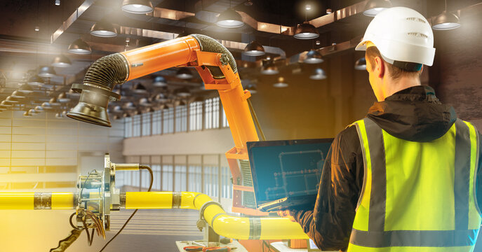 Industrial robotics, engineer monitors robotic arm and pipeline system via laptop, controlling automated processes and equipment setup in factory environment.