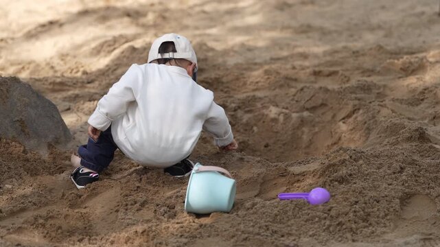 Little Asian Boy Squatting in a Sandbox and Digging Sand Outdoors.