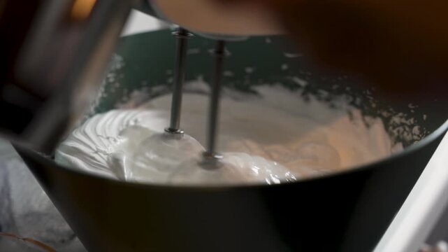 A thick white mixture is whipped with electric beaters in a deep bowl during a home baking process.