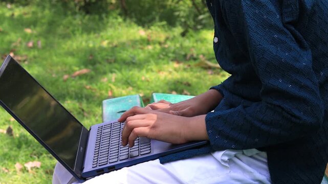 Teenager boy of Indian ethnicity sitting outdoor in nature and using laptop.