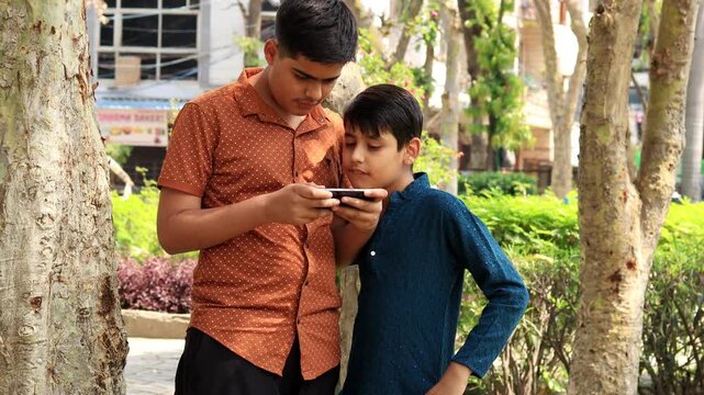 Two teenage children of Indian ethnicity standing outdoor in nature and using Mobile phone video portrait close up.