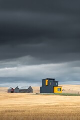 Fototapeta premium A contemporary house is located adjacent to a vintage barn, set against a backdrop of an expansive field beneath ominous dark clouds on a gloomy, overcast day in the countryside