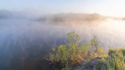 A serene morning view of a misty river at sunrise with lush green vegetation on the bank. © Александр Арендарь