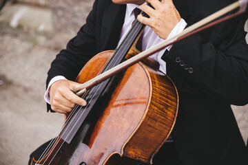 Fototapeta premium Close-up of a professional musician in a black suit playing a classic wooden cello with a bow at an outdoor event. Elegant string instrument performance capturing talent, culture, and formal music.