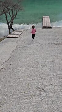 A young girl in a beige outfit joyfully runs toward a rustic wooden pier on the turquoise waters of Lake Kournas, Crete. A serene and picturesque travel moment. High quality FullHD footage