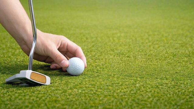 hand placing golf ball on green with putter, close up golf setup on putting green, preparing to putt on smooth turf, golfer aligning ball and club, sports equipment on grass.