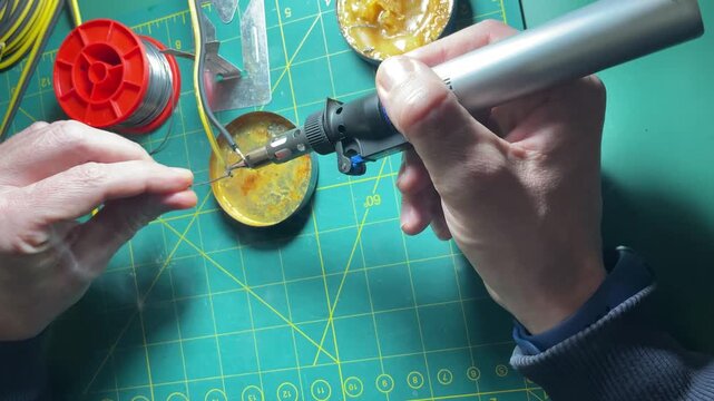 Close-up of a technician soldering electrical wires using a gas soldering iron, solder, and rosin. The video demonstrates tinning copper wires on a wooden workbench. High quality 4k footage