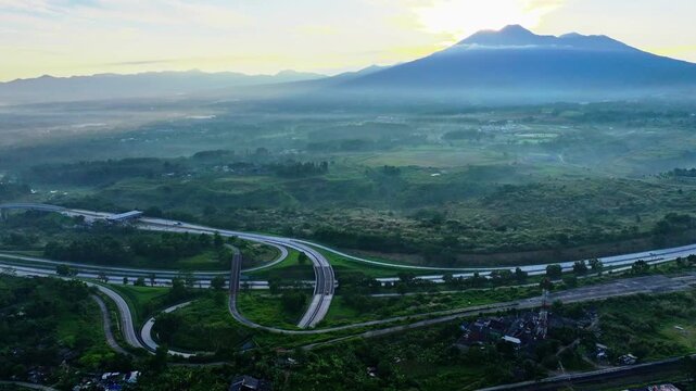 Aerial drone shot of a complex highway interchange winding through a lush green landscape at sunrise, with a majestic mountain and misty valley in the background.