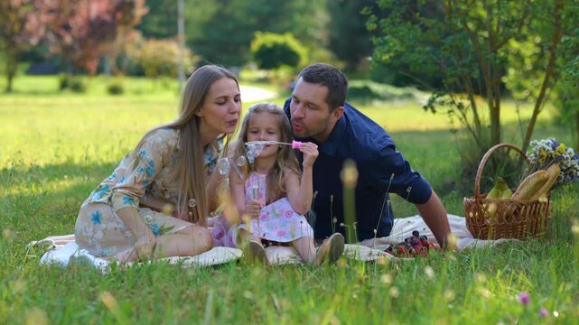 Young Caucasian parents spending weekend with child on picnic having fun blowing soap bubbles. Cheerful little girl with mother amd father on nature. Happy family outdoors. Slow motion