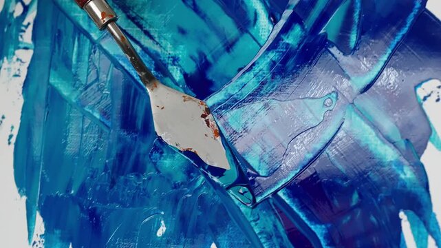 Close-up of palette knife spreading vivid blue and teal acrylic paint across white canvas, creating abstract impasto textures, layered strokes, and expressive marks in progress