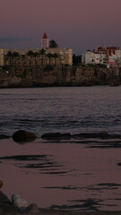 A tranquil coastal town at dusk with calm water reflecting the soft pink sky, rocky shoreline in the foreground, and silhouetted buildings and palm trees beyond. © Aperezer