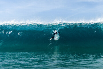Obraz premium A lone surfer duck dives beneath a towering turquoise wave, cutting through white spray as the ocean swells overhead under a clear, calm sky.