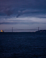 A long suspension bridge stretches across dark water at dusk, illuminated by small lights along its cables under a moody, cloudy evening sky. © Aperezer