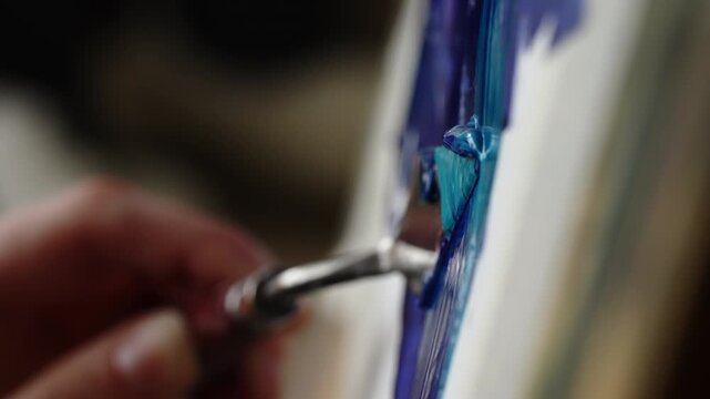 Cinematic close-up of an artist guiding a palette knife to sweep thick blue paint across a canvas, building luminous impasto textures and glossy layers during a studio session