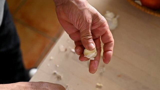 Close-up of a man's hands peeling a clove of garlic with a knife in the kitchen at home, detail of an organic ingredient for gourmet cooking.