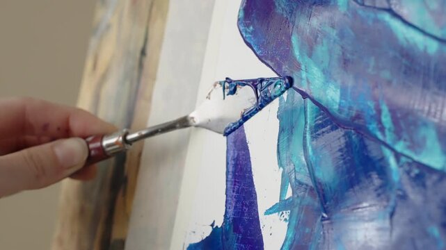 Close-up of a palette knife spreading thick blue and turquoise paint across a white canvas, creating textured abstract impasto strokes in a studio art painting process.