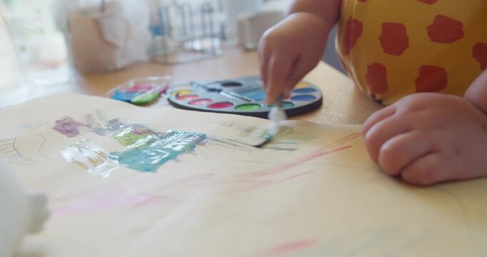 Unrecognizable toddler paints colorful watercolors on paper at a wooden table, focusing on early childhood education, creative expression, and fine motor skill development.