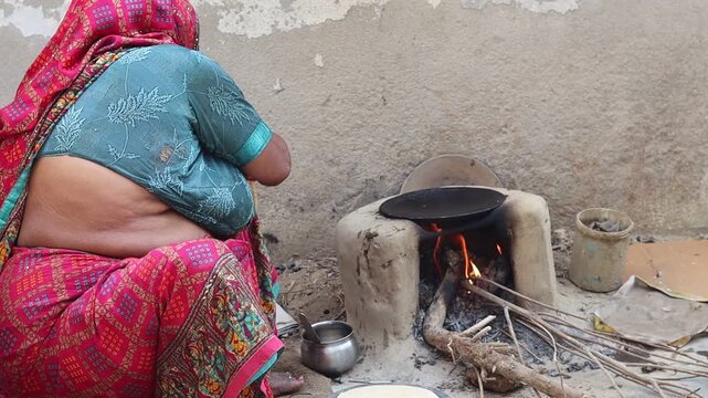 An elderly Indian village woman in traditional attire cooking handmade rotis (flatbread) on a traditional mud stove (chulha) using firewood in a rural kitchen.
