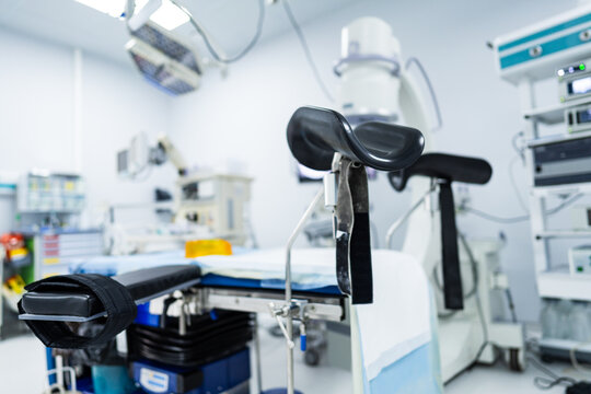 Surgical table in lithotomy position with medical imaging background. Modern operating theater with a patient table equipped with leg supports and stirrups for surgery.