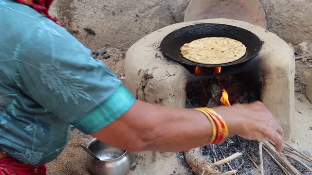 An elderly Indian village woman in traditional attire cooking handmade rotis (flatbread) on a traditional mud stove (chulha) using firewood in a rural kitchen.
