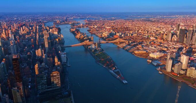 Footage above the East River approaching the Queensboro Bridge. Cityscape of Queens is lit by the setting sun.