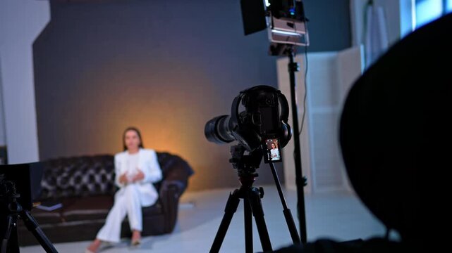 Filming equipment is directed on the woman in white suit in the blurred backdrop. Lady talks to camera gesturing. Blog footage.