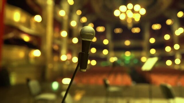 Grand theater stage with microphone in foreground, opulent chandeliers and balcony seating bathed in golden lights, empty venue ready for symphony or gala