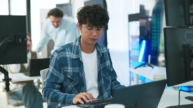 focused young male developer with curly hair in blue plaid shirt intensely codes on laptop in modern AI office, surrounded by multiple monitors displaying code neural networks and data graphs	