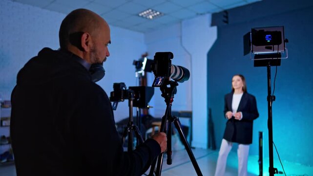 Male photographer in headphones and hoodie holds a camera and looks at display. Lady in black jacket is talking to camera.