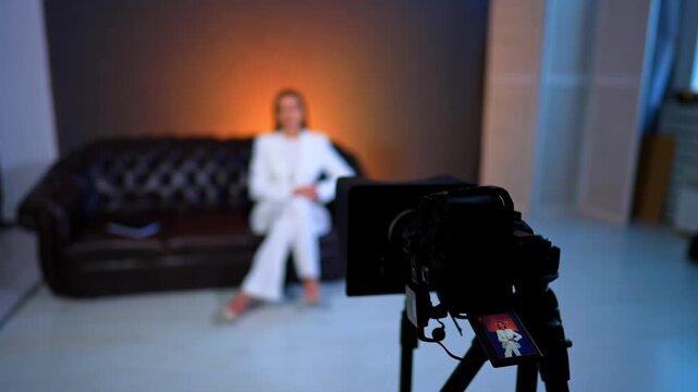 Camera is filming a woman sitting and speaking on the sofa. Blurred backdrop. Footage backstage.