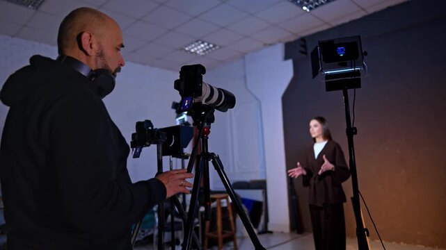 Caucasian cameraman looks focused at camera display. Photographer records a long-haired woman standing at blurred backdrop.