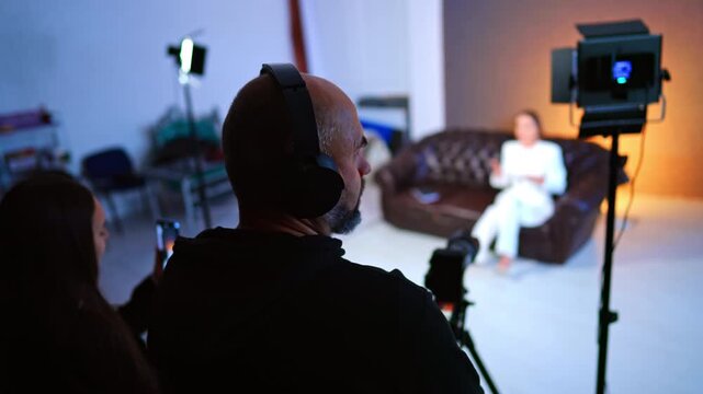 Rear view of the male photographer wearing headphones standing near the camera. A woman in white suit sits on the sofa talking for blog.