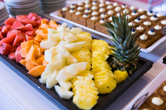 Fresh fruit platter with sliced watermelon, cantaloupe, honeydew, and pineapple on a black tray, alongside gourmet dessert squares topped with cream and blueberries at a bright catered buffet.