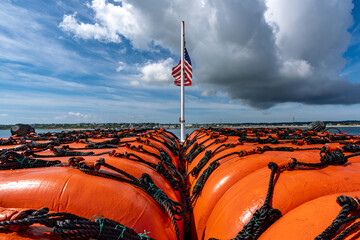 A row of bright orange life rafts tied down with black netting sits in the foreground, looking toward a flagpole flying the American flag against a cloudy blue sky. 