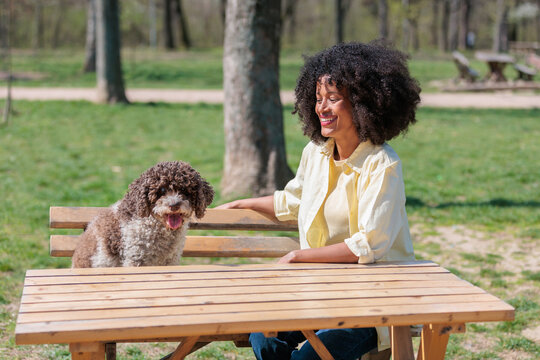 Woman sits at a table in a park with her dog enjoying a sunny day outdoors during spring time with trees in the background